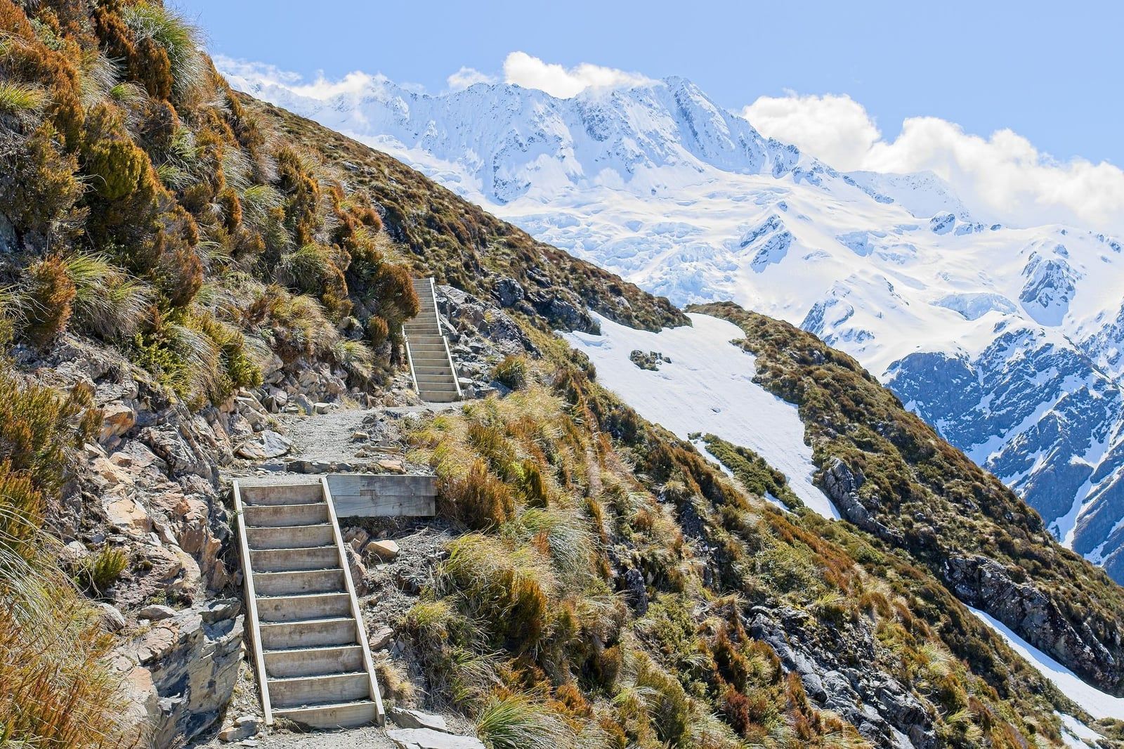 Wooden steps climb a steep alpine slope on the Sealy Tarns Track opposite snow-capped peaks in Aoraki Mount Cook National Park.
