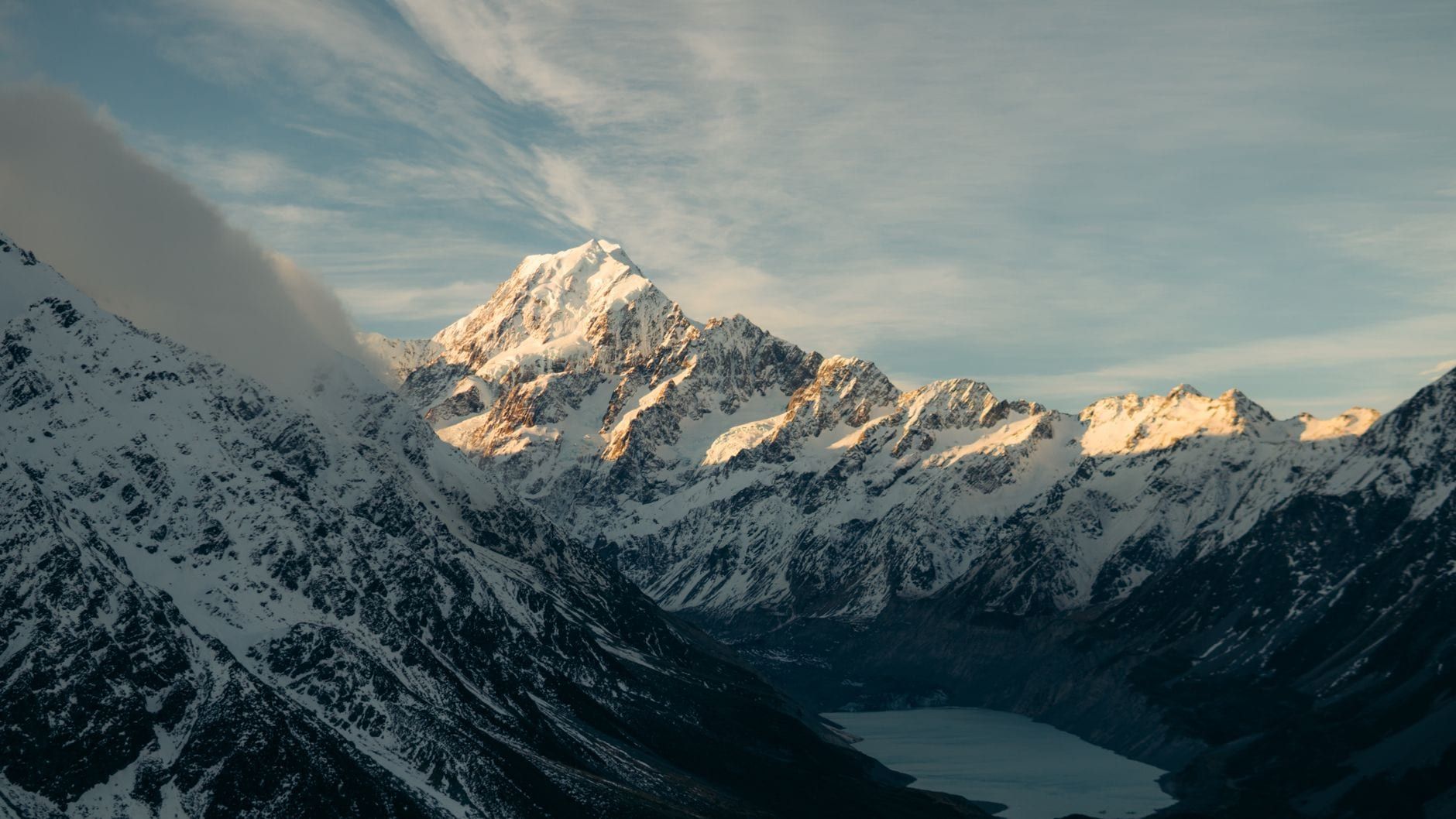Snow-covered peak of Mount Cook glowing in morning sunlight above a deep alpine valley and glacial lake.