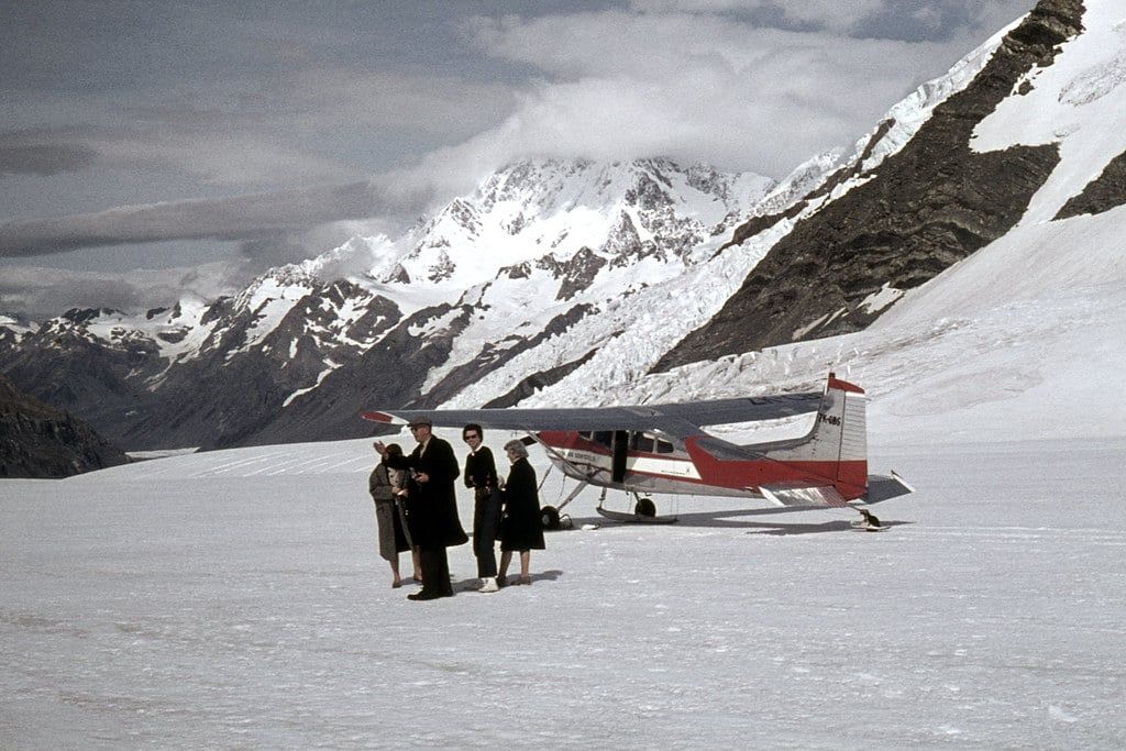 A red and white ski plane rests on the snow of the Tasman Glacier near Mount Cook with passengers standing beside it.