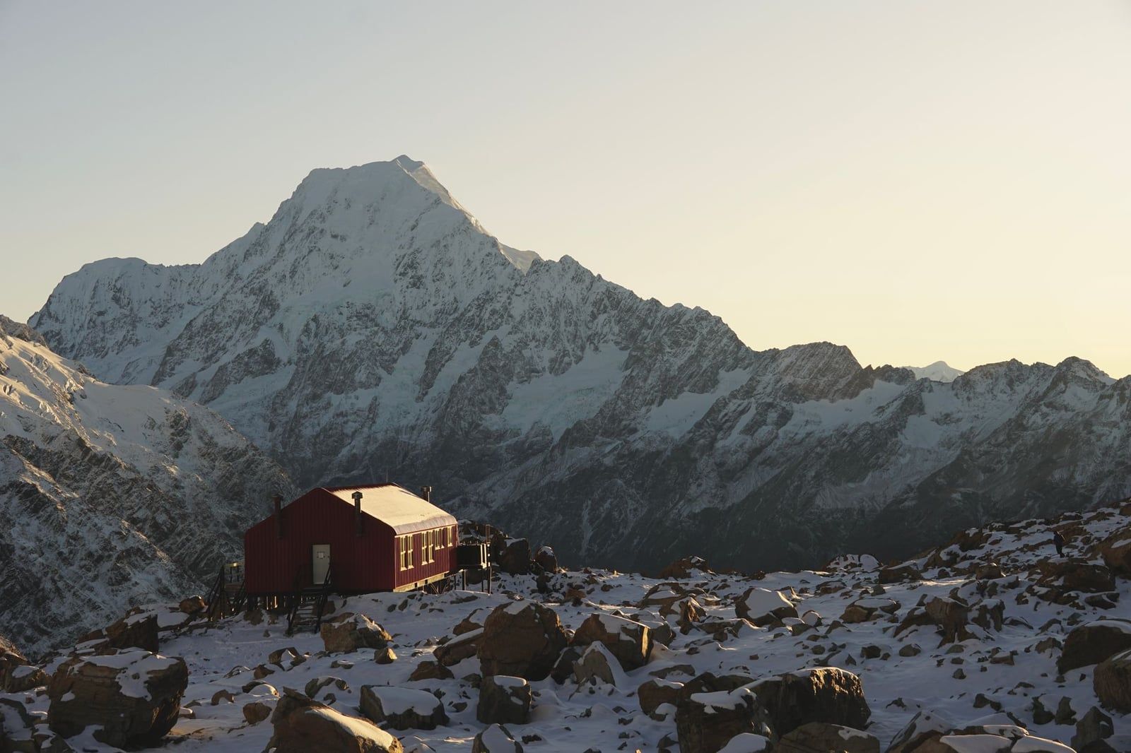 A red alpine hut sits on snow-dusted rocky terrain with the snow-covered peak of Mount Cook rising behind it.