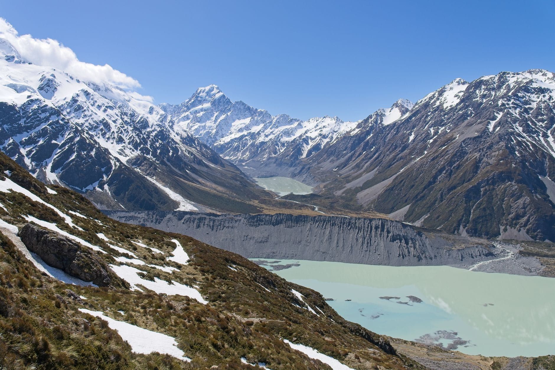 Wide panoramic view of snow-dusted mountains and glacial lakes with Mount Cook rising in the distance under a clear blue sky.