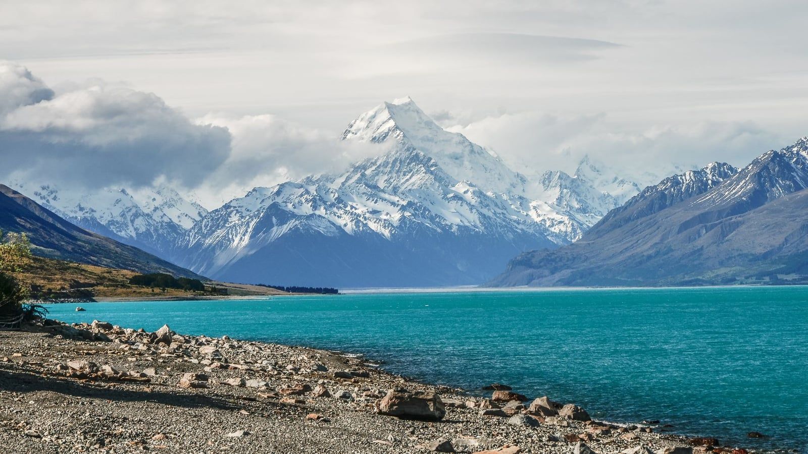 Turquoise waters of Lake Pukaki with a snow-covered Mount Cook rising in the distance under a cloudy sky.